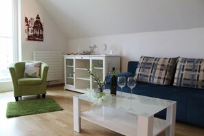 Ein Wohnzimmer mit einem grünen Sessel, einem weißen Couchtisch mit zwei Weingläsern, einem blauen Sofa mit karierten Kissen und einer weißen Kommode mit Glastüren.