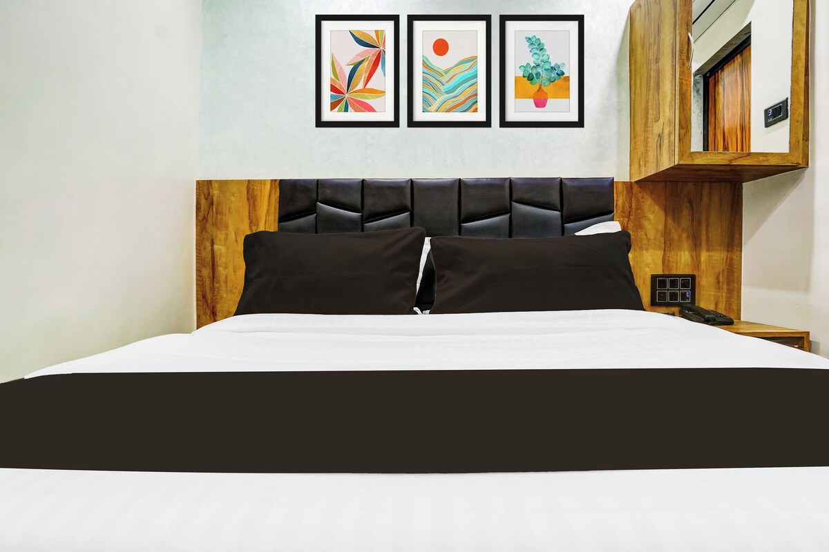 Una cama con cabecera negra y ropa de cama blanca, una mesita de noche de madera con un teléfono, y tres obras de arte enmarcadas en la pared.