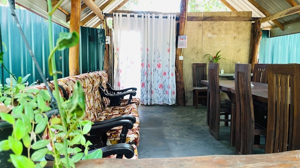 A covered outdoor seating area with a sofa and wooden chairs, a table, and a view through a curtain.