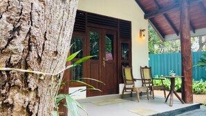 Una puerta de madera con un ventanal, un patio techado con sillas y una mesa, y un árbol en primer plano.