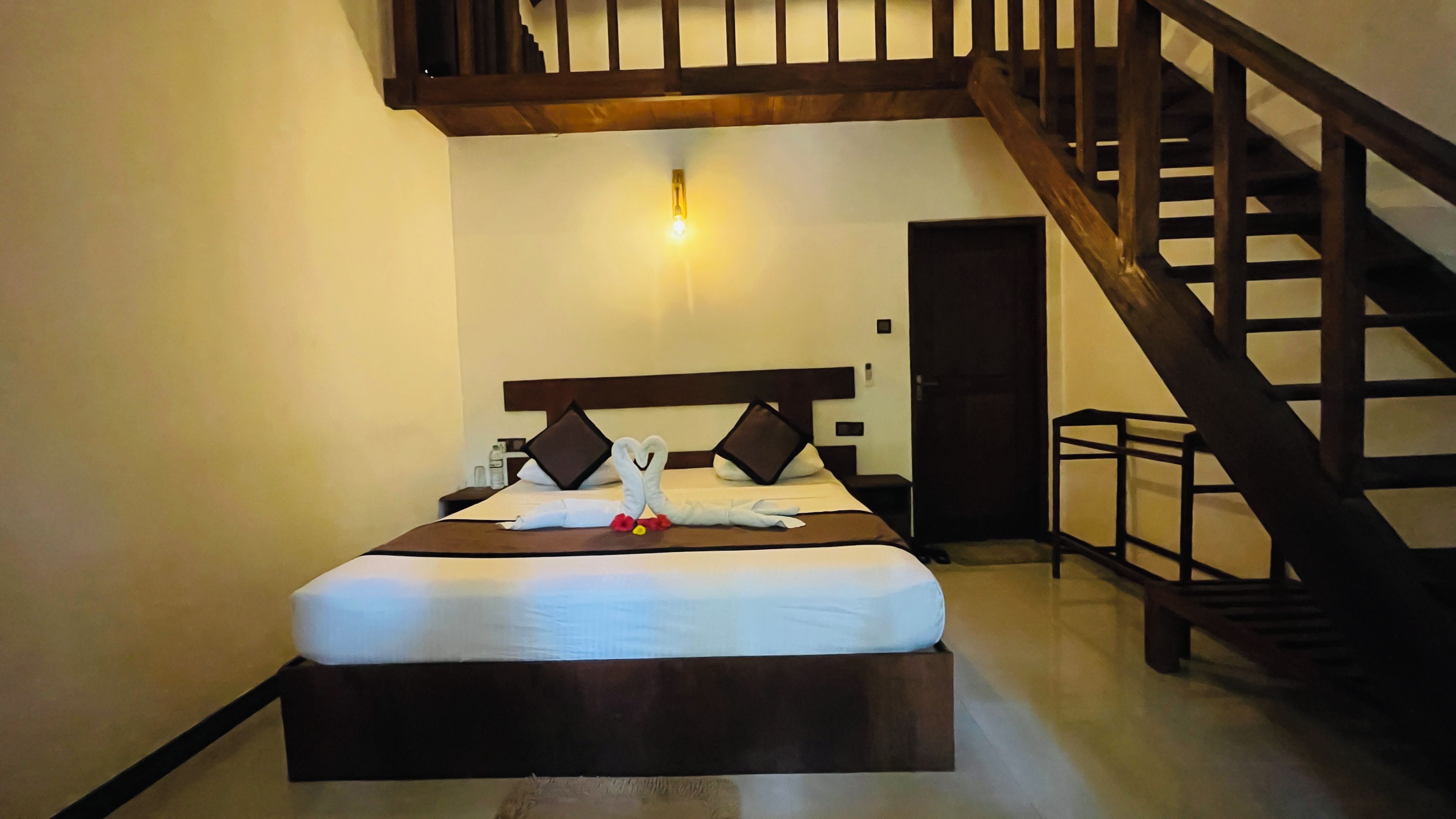 A hotel room with a bed, a wooden headboard, a nightstand, a wall-mounted light, and a staircase.