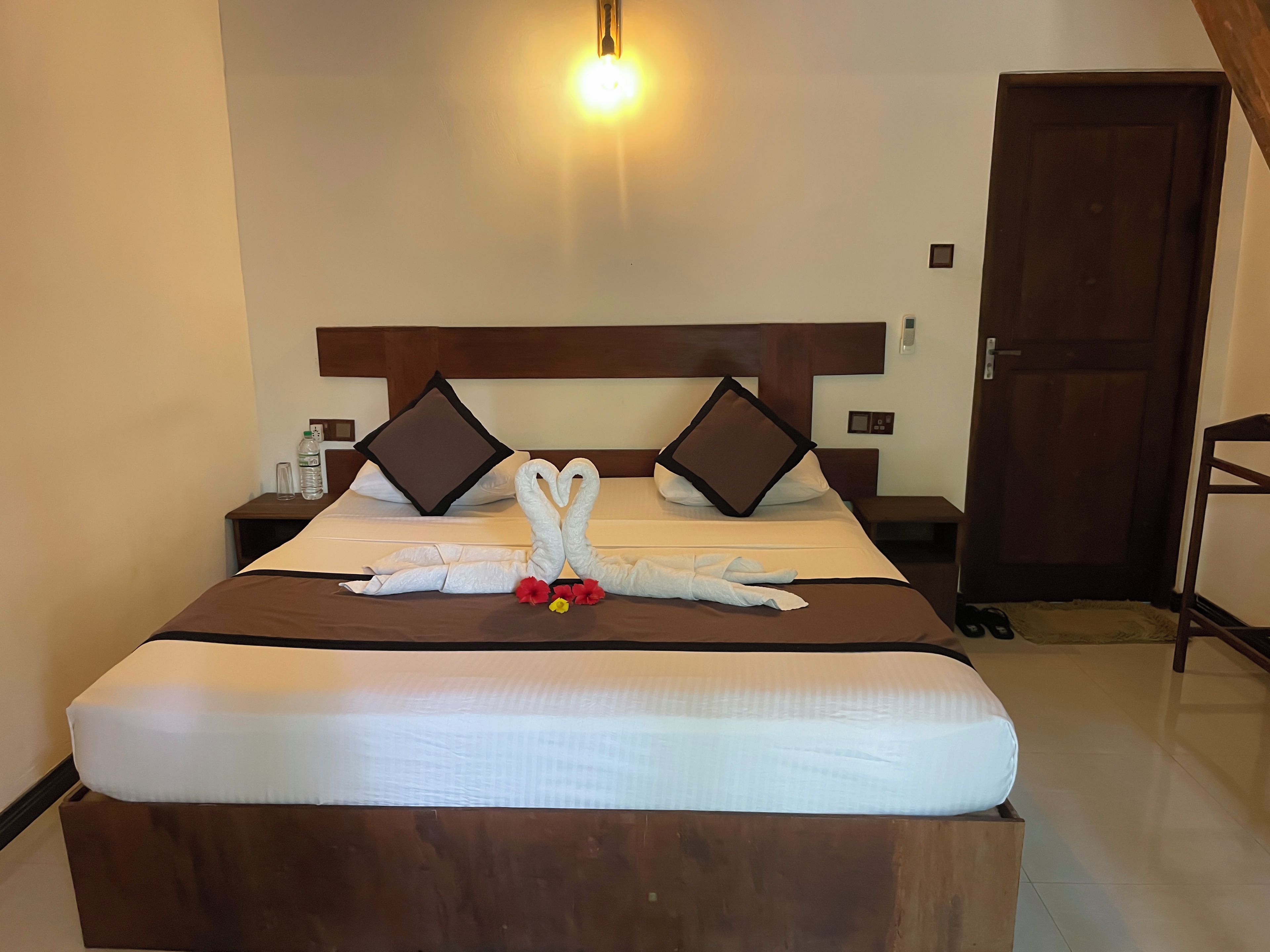 A hotel room with a bed, two pillows, a bedside table with a bottle, and a door.