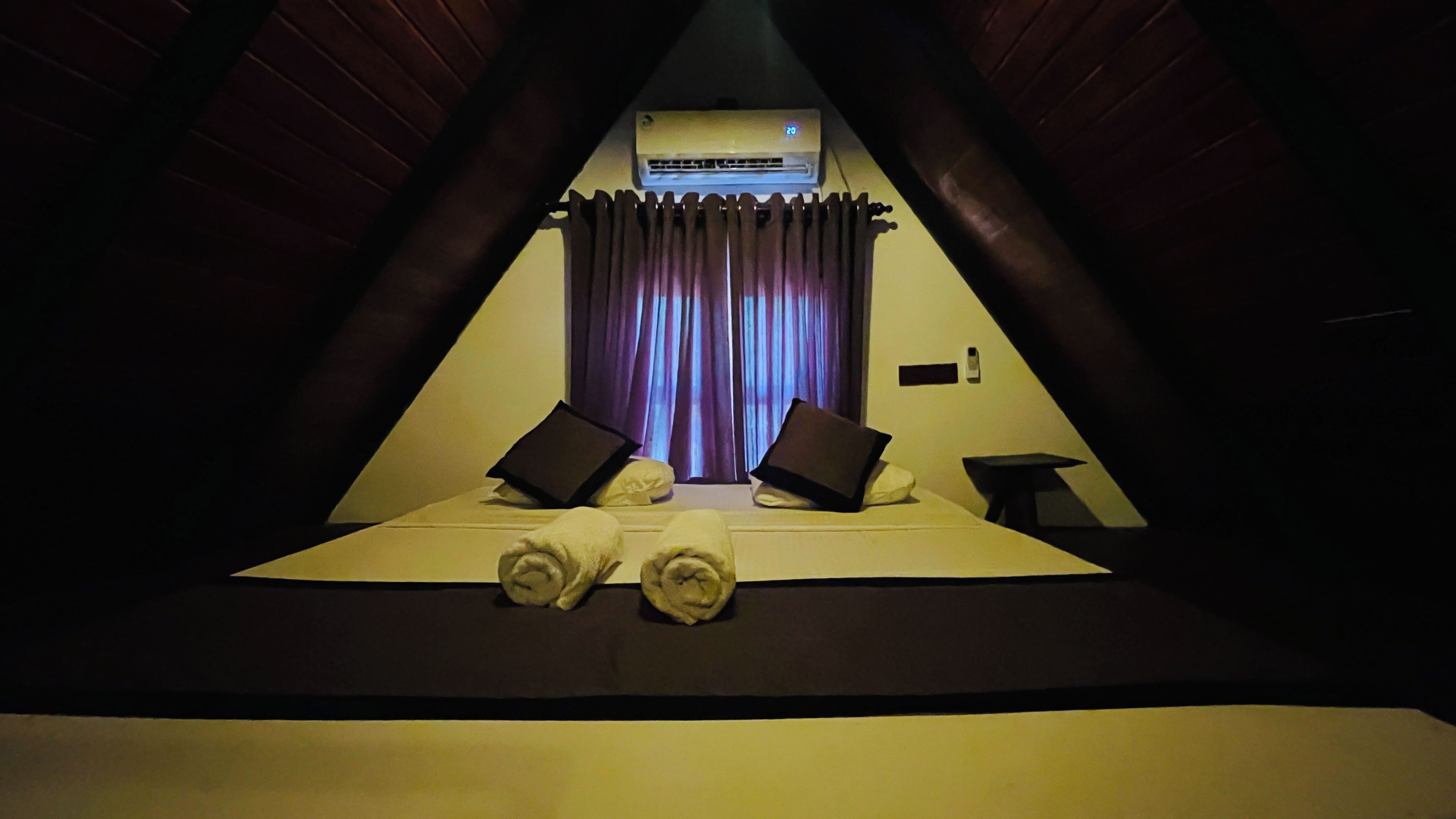 A triangular room with a bed, two pillows, a small table, and an air conditioning unit.