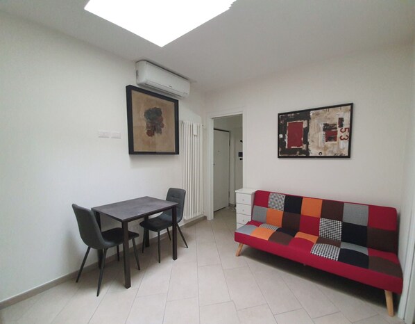 Quarto com mesa de jantar e cadeiras, sofá colorido, aparelho de ar condicionado e duas obras de arte emolduradas nas paredes.