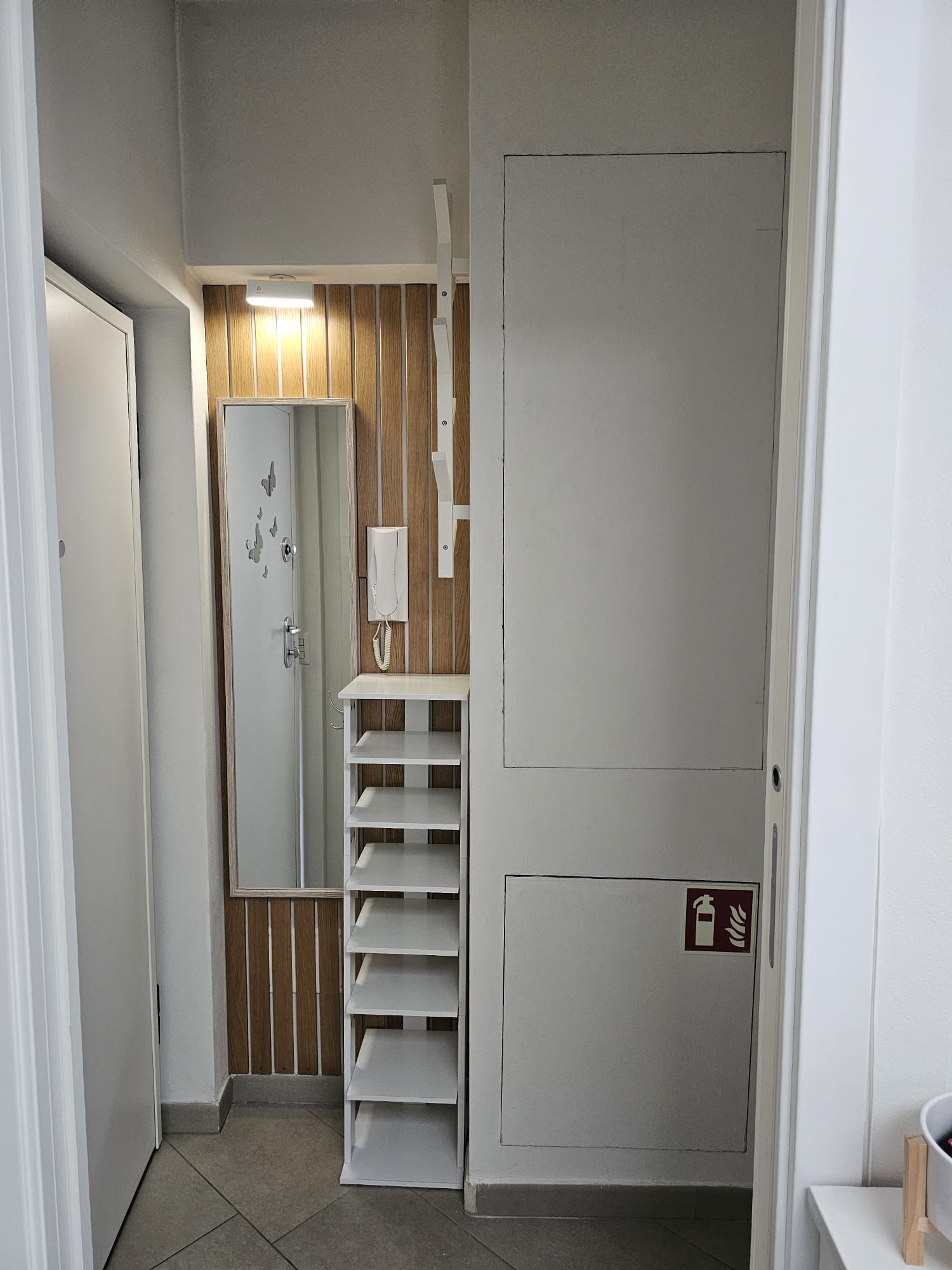 A hallway with a white door, a wooden panel wall, a white shelf unit, and a fire safety sign.