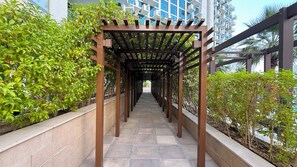 A covered walkway with a pergola structure, flanked by greenery and a modern building in the background. - Azizi Signature Studio Al Jaddaf (Dubai)