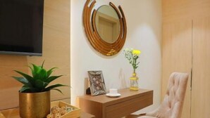 A modern living room with a flat-screen TV, a round mirror, a wooden sideboard, a cushioned armchair, and a potted plant. - Hotel Adora (New Delhi)