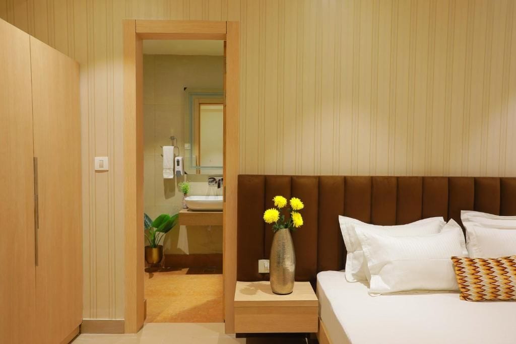 A hotel room with a bed, a bedside table with a vase of flowers, a wooden wardrobe, and a bathroom visible through an open door.
