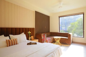 Ein modernes Hotelzimmer mit einem großen Bett, einer Couch, einem kleinen runden Tisch und einem großen Fenster mit Blick auf Grünflächen.