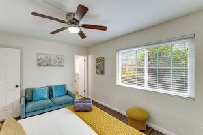 Ein Schlafzimmer mit einem Bett, einem blauen Sofa, einem Deckenventilator, einem Fenster mit Jalousien und einem kleinen runden Hocker.