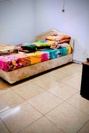 Una cama con colcha estampada y varias almohadas, suelo de baldosas y pared lisa con toma de corriente.