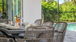 Área de refeições ao ar livre com cadeiras de vime e mesa posta para uma refeição, com vista para uma piscina e vegetação exuberante.