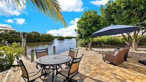 A patio with a table and chairs, overlooking a body of water, with a palm tree and greenery in the background. - Kayak + Fishing Dock | Pool + Spa | Mangroves + Beaches | Sanctuary Point (Bonita Springs)