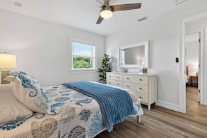 Un dormitorio con cama, cómoda, ventilador de techo, ventana y puerta que da a otra habitación.