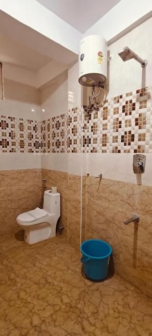 A bathroom with a toilet, a shower, and a blue bucket on the floor. - Roja Palace (Mahabalipuram)