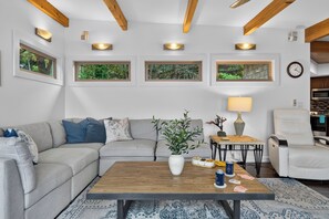 Een moderne woonkamer met een hoekbank, een salontafel, een bijzettafeltje met een lamp, en uitzicht op het landschap door ramen.