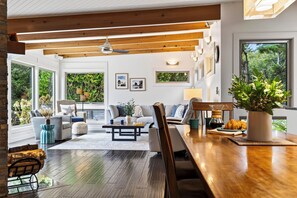 A modern living room with a wooden ceiling, large windows, and a dining area with a wooden table and chairs. - Ocean-View Deck + Fire Pit + BBQ | Short Walk to the Beach | The Grey Piper (Arch Cape)