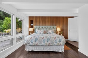 A bedroom with a large bed, wooden panel walls, a view of greenery outside, and a white door. - Ocean-View Deck + Fire Pit + BBQ | Short Walk to the Beach | The Grey Piper (Arch Cape)