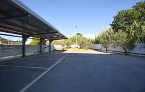 Een overdekte parkeerplaats buiten, met een heldere lucht, bomen en een hek.