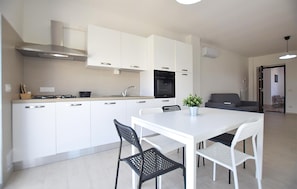 Een moderne keuken met witte kastjes, een fornuis en een oven. Er is een eettafel met twee stoelen en een bloempot.
