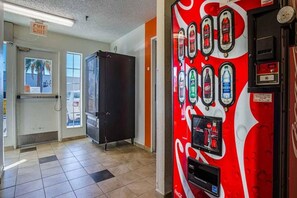 A vending machine with various drink options, a door with an exit sign, and a window with a view of palm trees. - Motel 6 Tucson, Az - North_2 Full Beds | (Tucson)