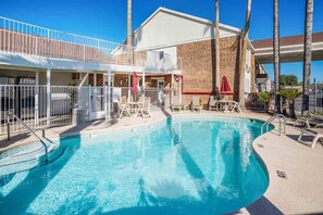 A swimming pool with a clear blue water, surrounded by lounge chairs and umbrellas, and a building with a brick facade in the background. - Motel 6 Tucson, Az - North_2 Full Beds | (Tucson)