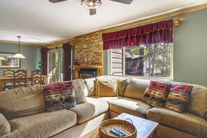 A cozy living room with a stone fireplace, a large window with red curtains, and a woven sofa with patterned cushions. - Walk to Ski & Main St! Newly Refinished w/ Hot Tub (Breckenridge)