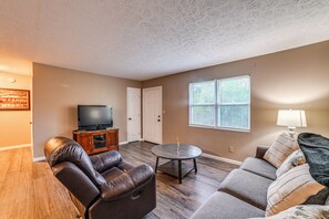 A living room with a leather couch, a small round table, a television on a wooden stand, and a window with blinds.