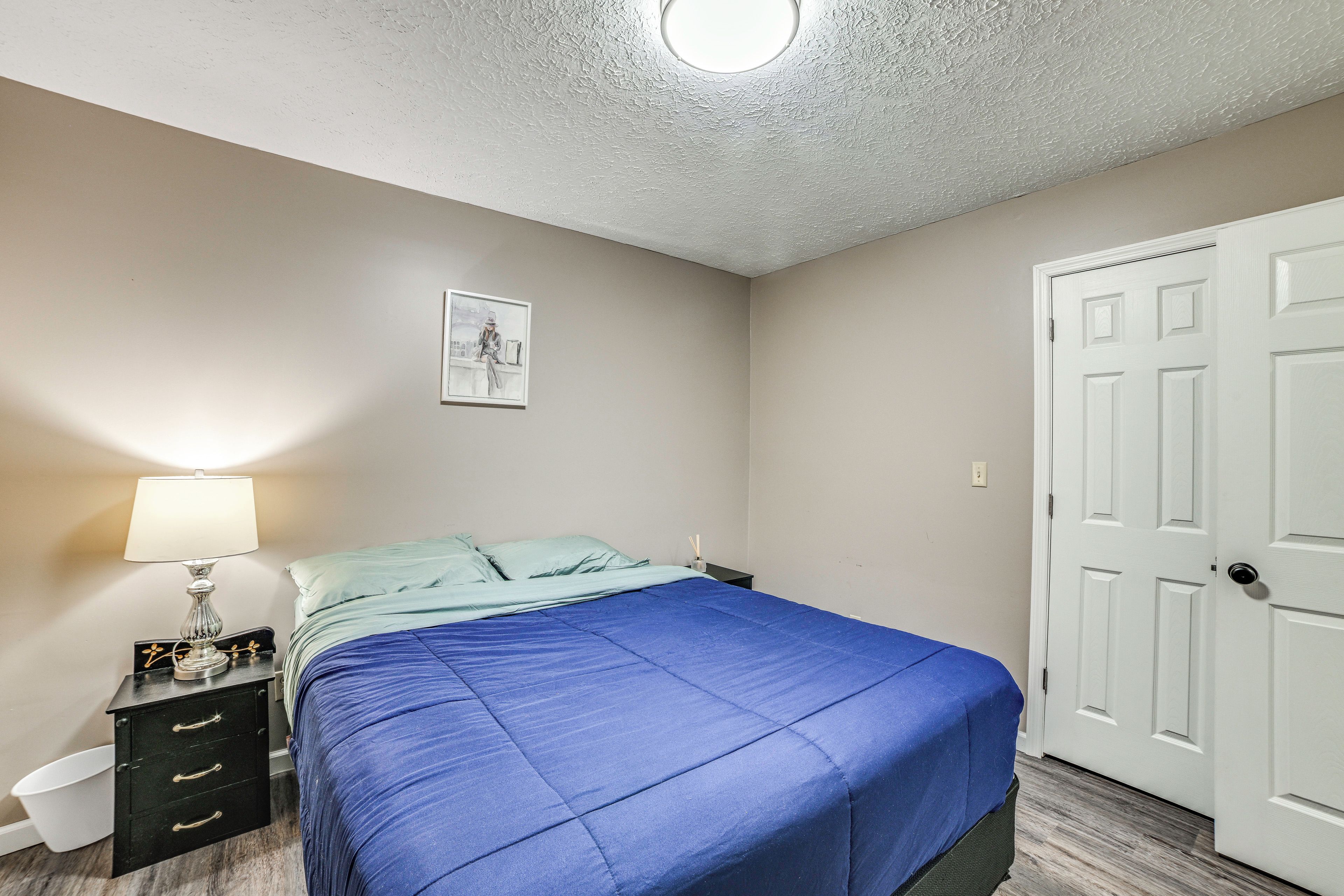 Un dormitorio con cama, mesita de noche, lámpara, puerta y un cuadro colgado en la pared.