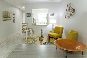 A living room with a sofa, armchair, and a unique orange chair. There is a patterned rug, a small table with a candle, and a lamp. - Gulf Coast Corner Condo Near Charlotte Harbor (Punta Gorda)