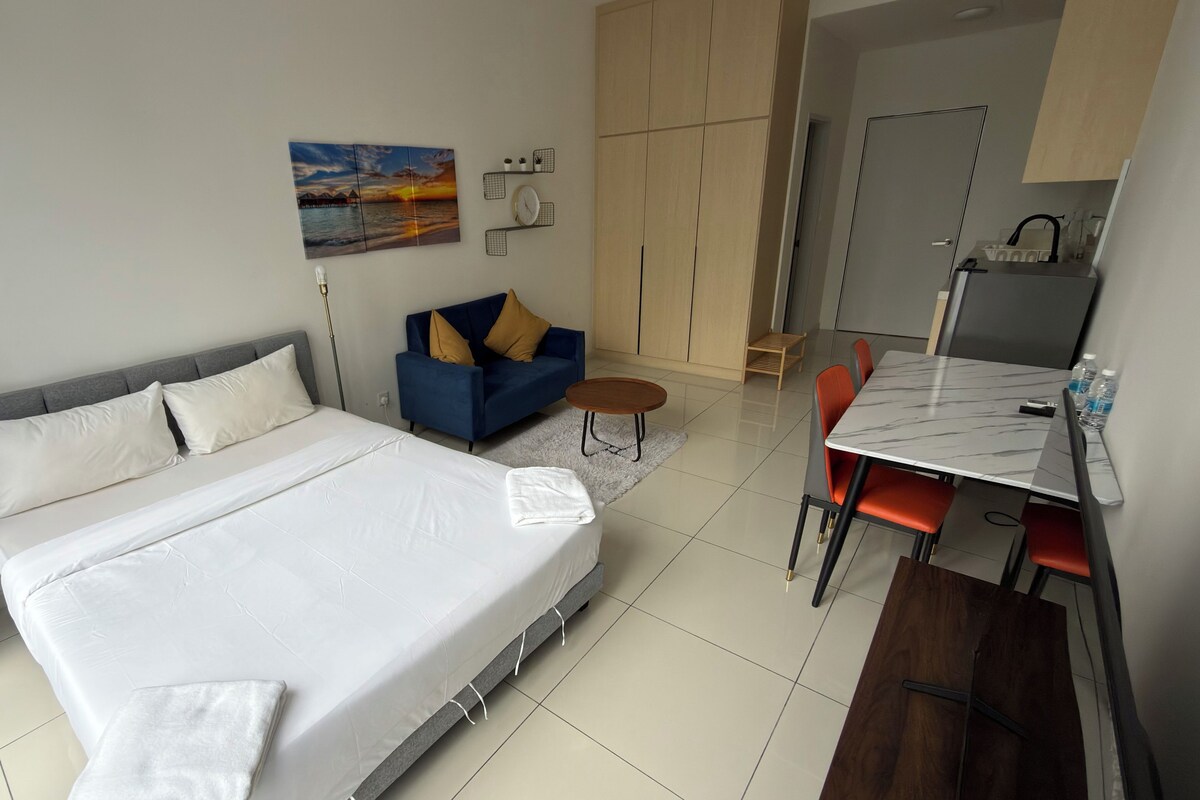 Habitación de hotel con cama, sofá, mesa de comedor con sillas, una mesita auxiliar y un cuadro en la pared.
