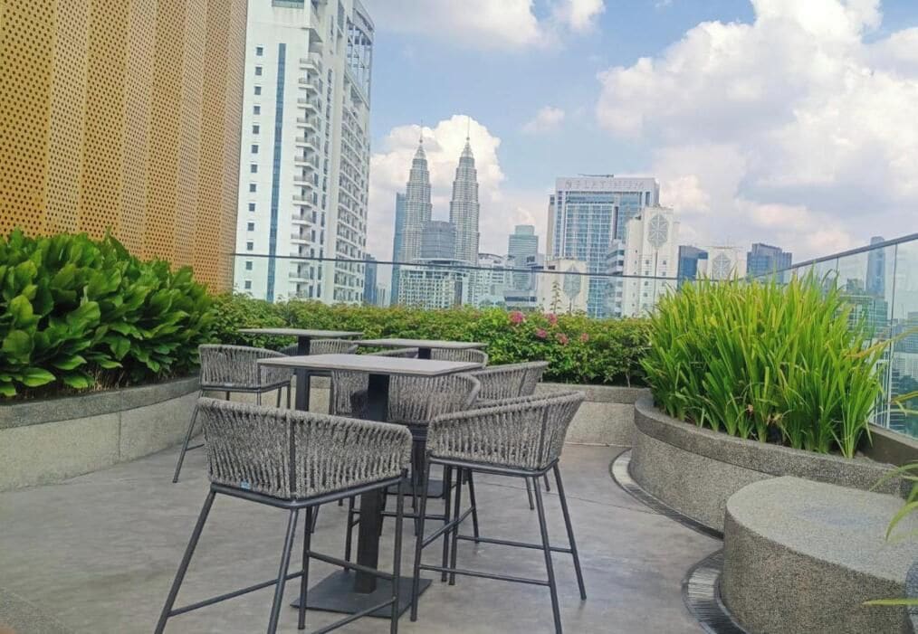 Un espacio al aire libre con mesas y sillas, rodeado de follaje, con vista a un paisaje urbano de edificios altos.