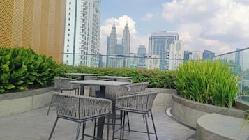 Un espacio al aire libre con mesas y sillas, rodeado de follaje, con vista a un paisaje urbano de edificios altos.