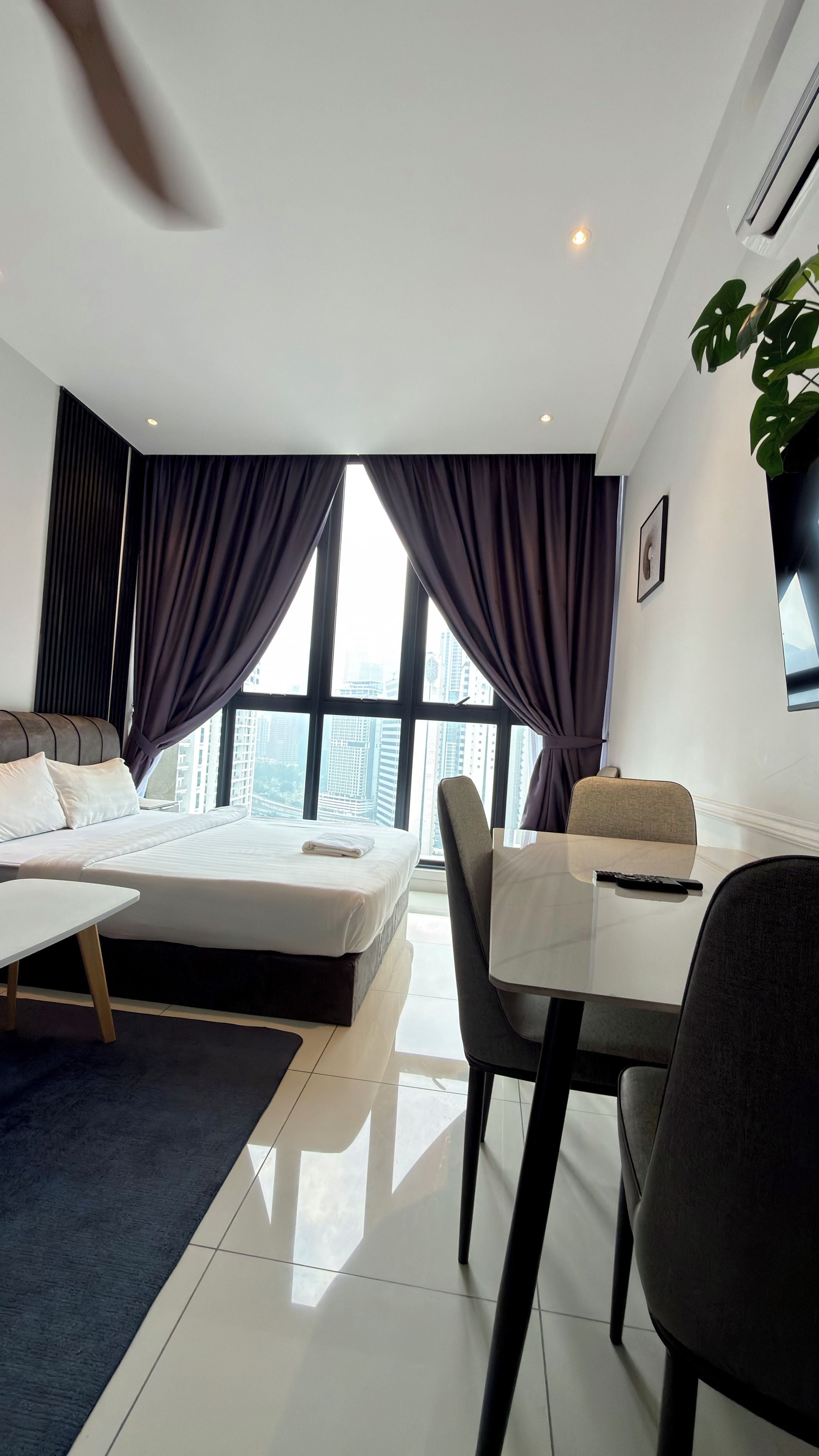 Habitación de hotel moderna con cama, una mesa de comedor y sillas. Cuenta con un ventanal con cortinas, una planta y un cuadro en la pared.