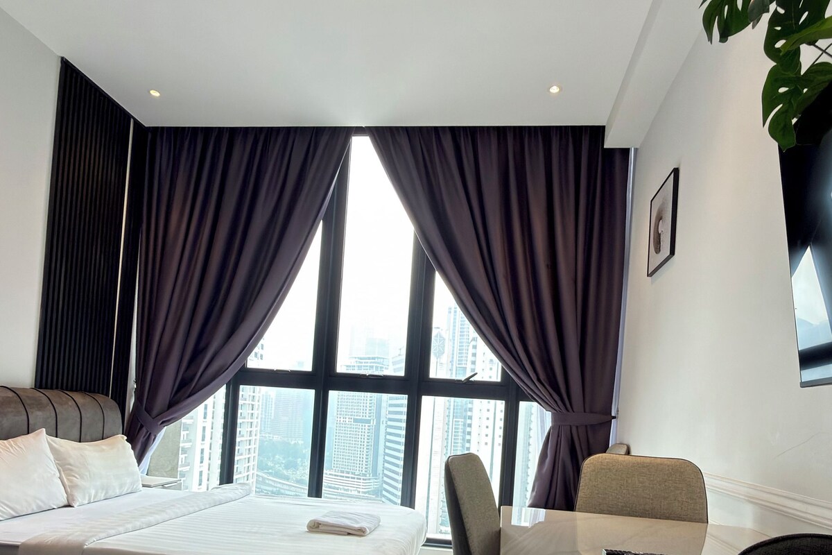 Habitación de hotel moderna con cama, una mesa de comedor y sillas. Cuenta con un ventanal con cortinas, una planta y un cuadro en la pared.
