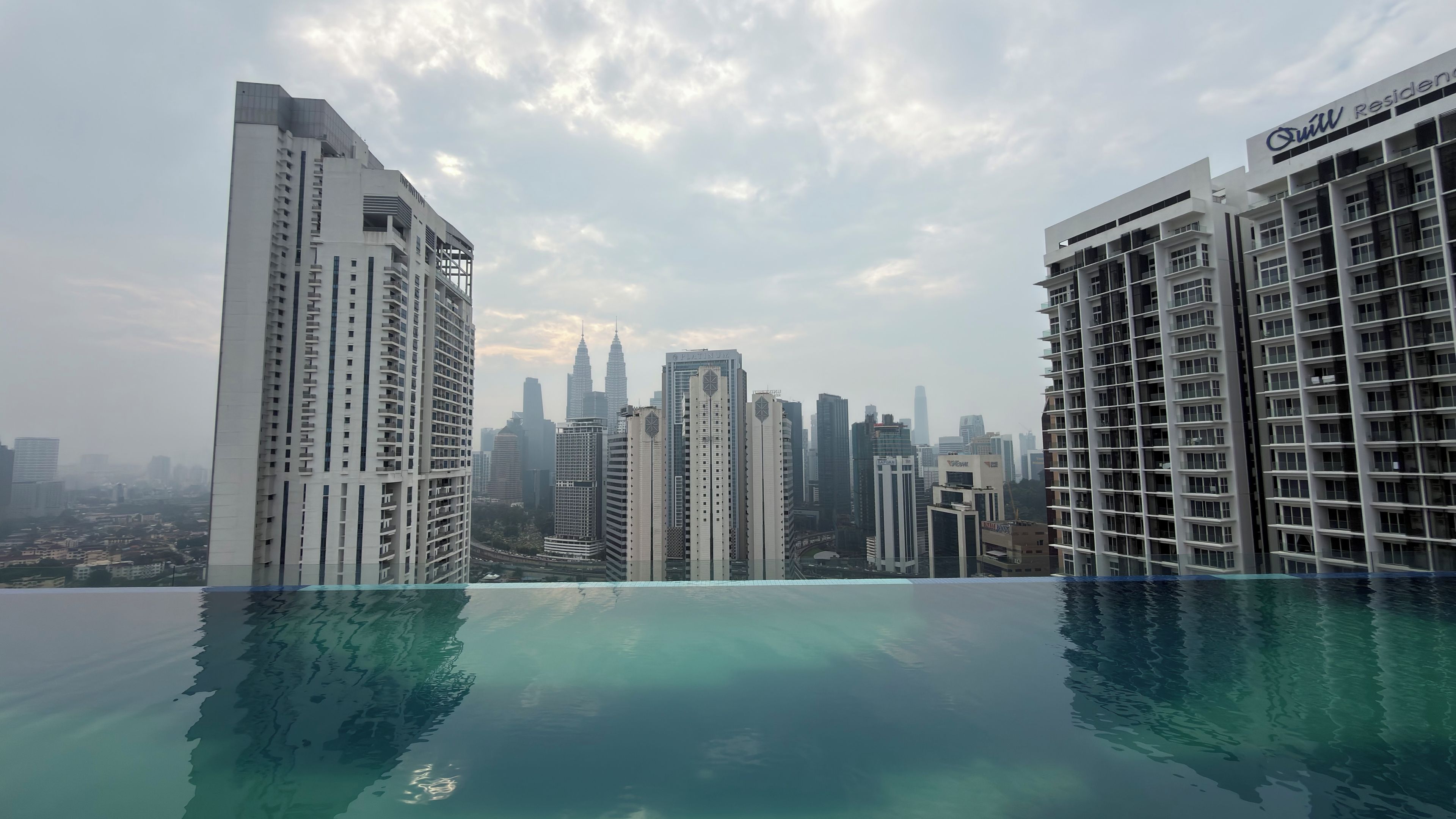 Un edificio de gran altura con vistas despejadas al horizonte de la ciudad, incluyendo las Torres Petronas, y una piscina con superficie de agua azul cristalina.