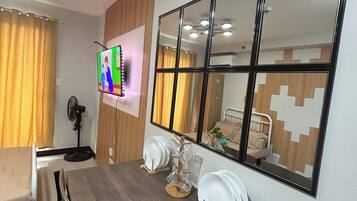 A dining area with a wooden table, chairs, a TV mounted on the wall, and a large mirror reflecting the room.