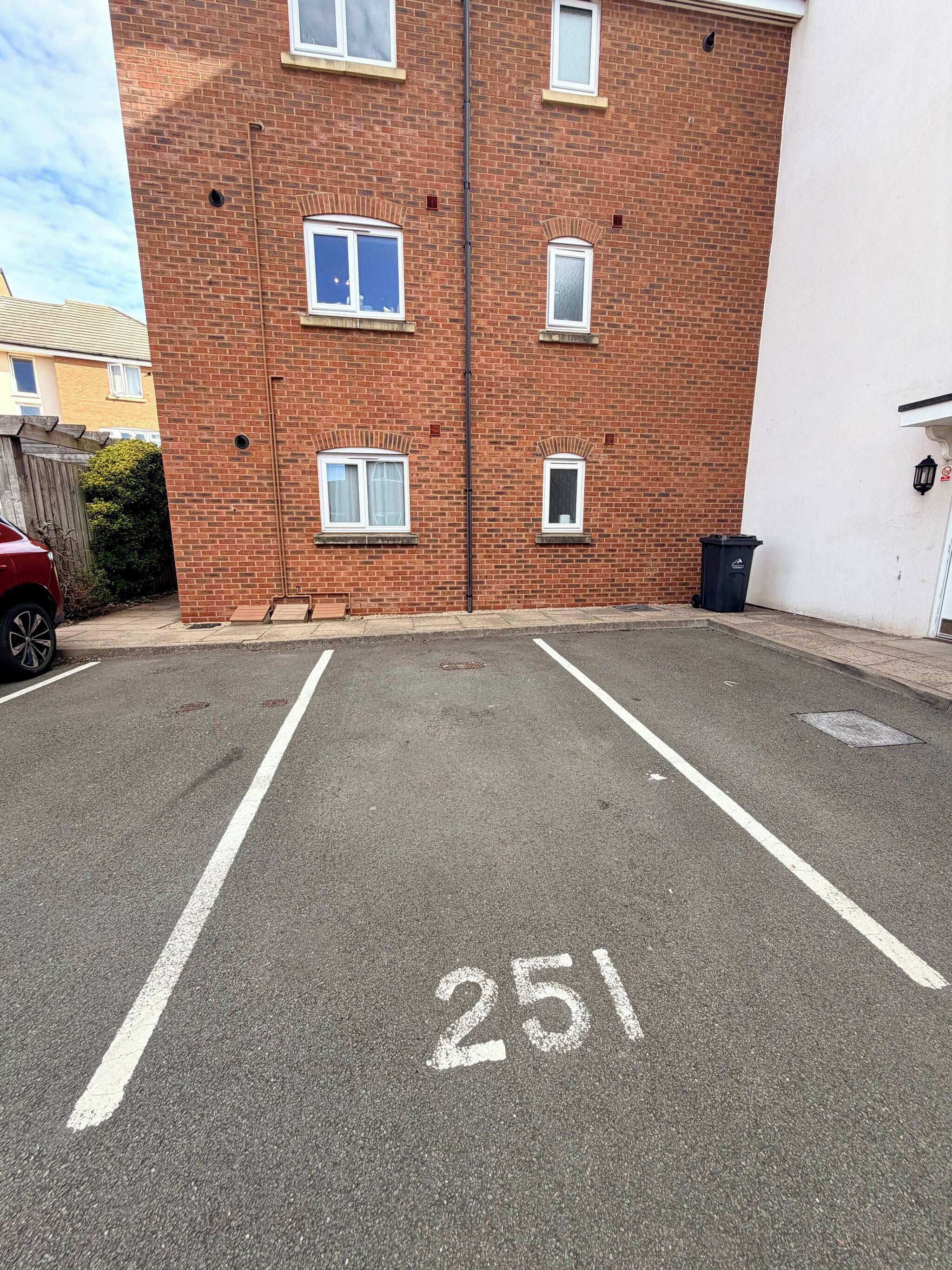 Un espace de stationnement avec un bâtiment en briques en arrière-plan, une voiture rouge garée et une poubelle sur le côté.
