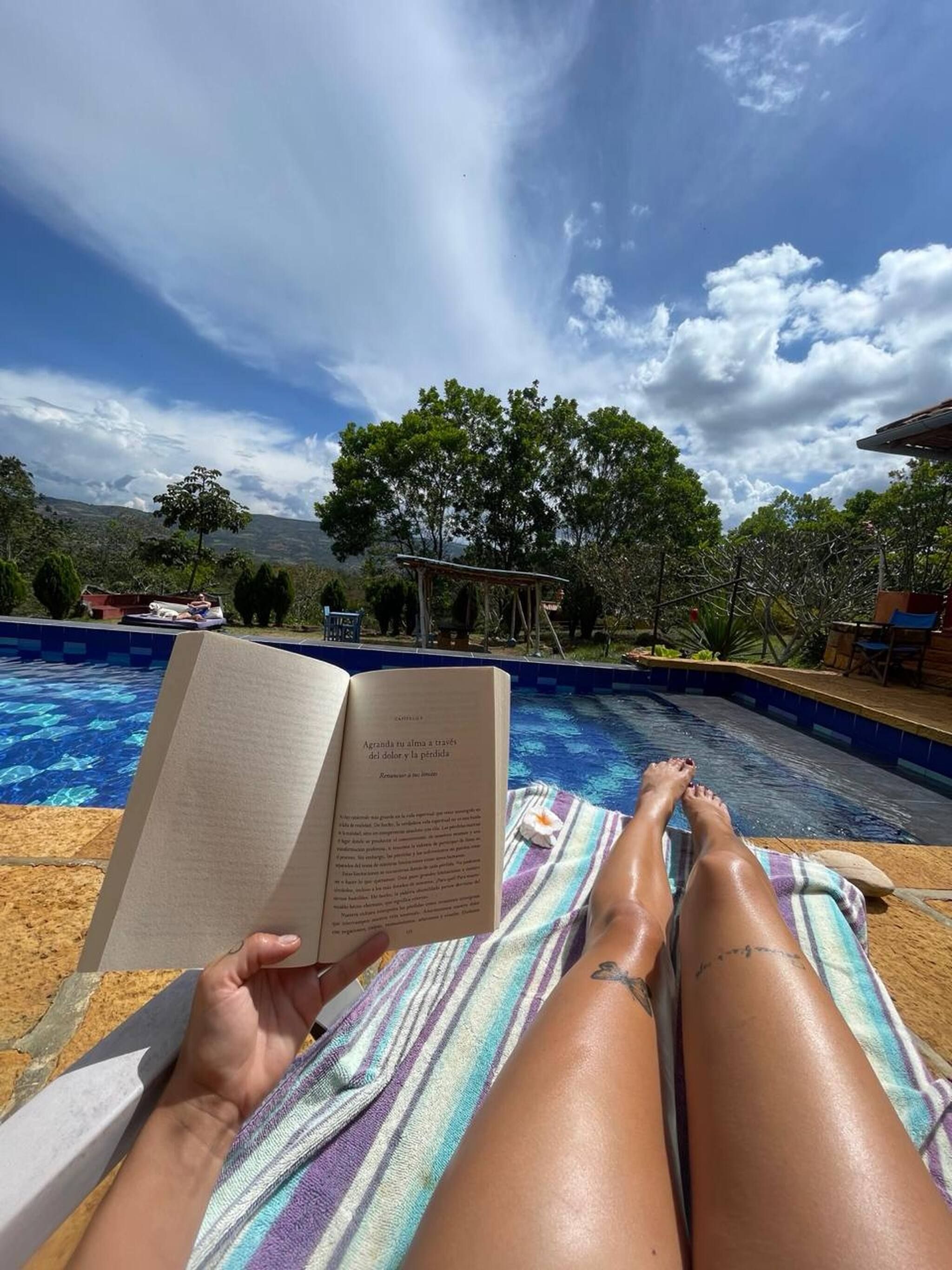 A person relaxing by a poolside with a book, surrounded by greenery and a clear sky.