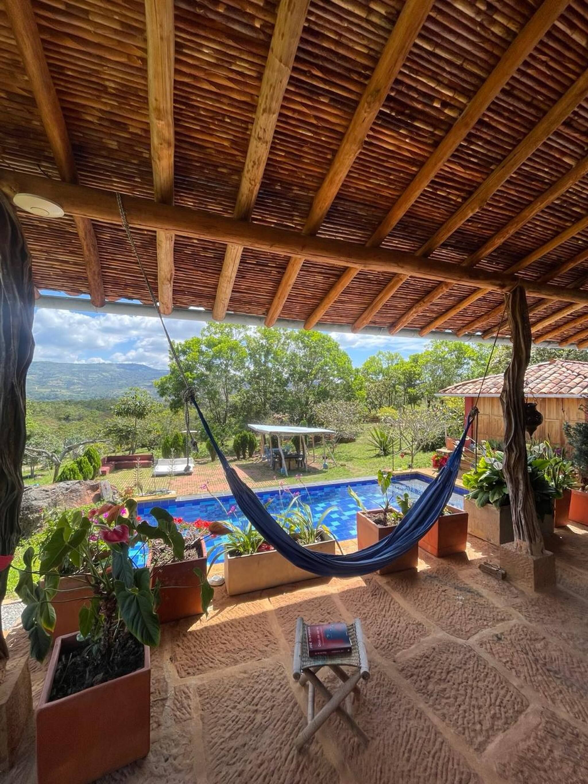 A hammock under a wooden roof, a small table with a book, and a pool with potted plants around it.