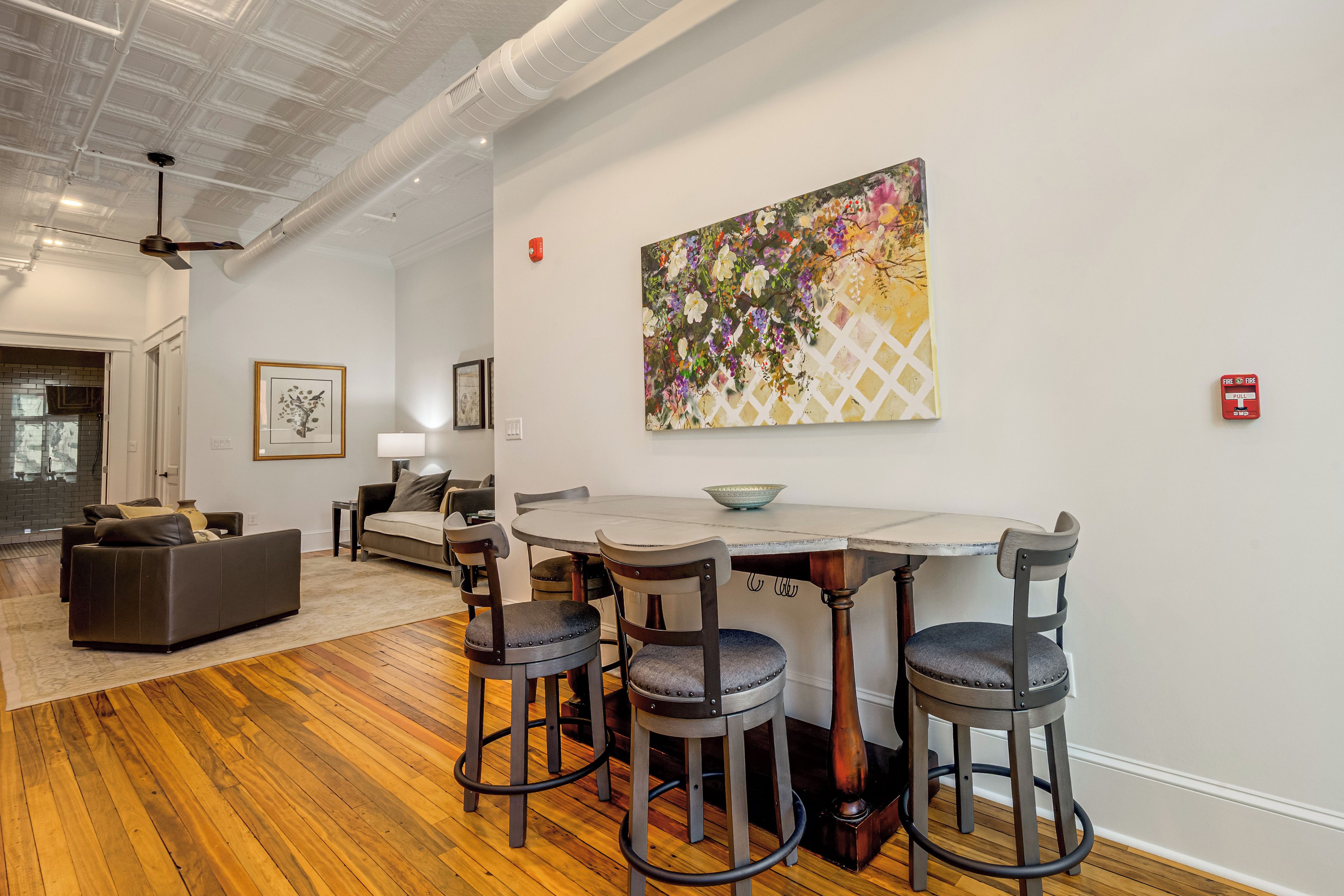 A modern interior with a dining area featuring a white table and four bar stools, a living room with a sofa and a fireplace, and a large floral painting on the wall.