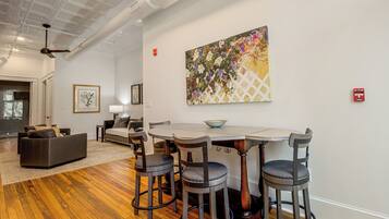 A modern interior with a dining area featuring a white table and four bar stools, a living room with a sofa and a fireplace, and a large floral painting on the wall.