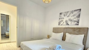 Un letto rifatto con lenzuola bianche e un asciugamano arrotolato, un pavimento in legno, un quadro astratto appeso al muro e una lampada a sospensione.