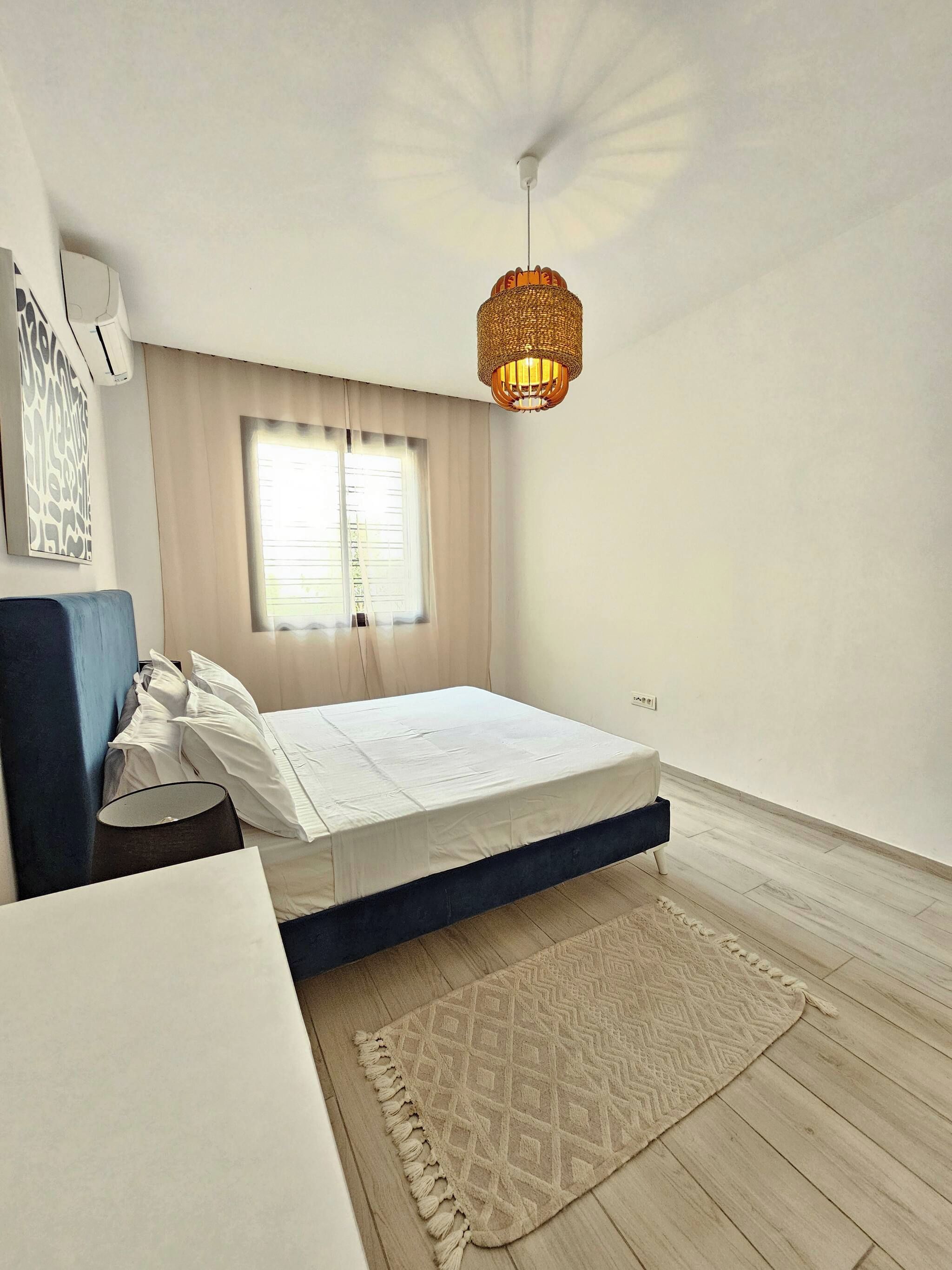 Ein Schlafzimmer mit einem Bett, einem Nachttisch, einer Lampe, einer Hängelampe und einem Fenster mit Vorhängen.