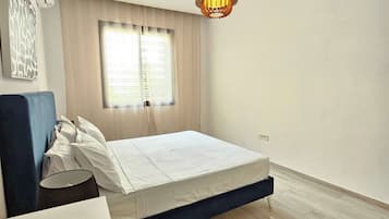 Quarto com cama, mesa de cabeceira, luminária, lustre e janela com cortinas.
