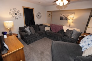 A living room with a sectional sofa, a wooden side table, a lamp, and a ceiling fan.