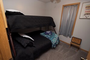 A single bed with a patterned bedspread, a pillow, and a blanket. There is a small wooden stool with a sign on it. A window with curtains is visible.