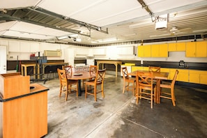 A spacious industrial-style room with a large table and chairs, a kitchen area with yellow cabinets, and a ceiling with exposed beams and lighting fixtures.