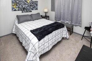 Um quarto com cama, mesa de cabeceira, luminária, janela com cortinas e uma parede com obras de arte.