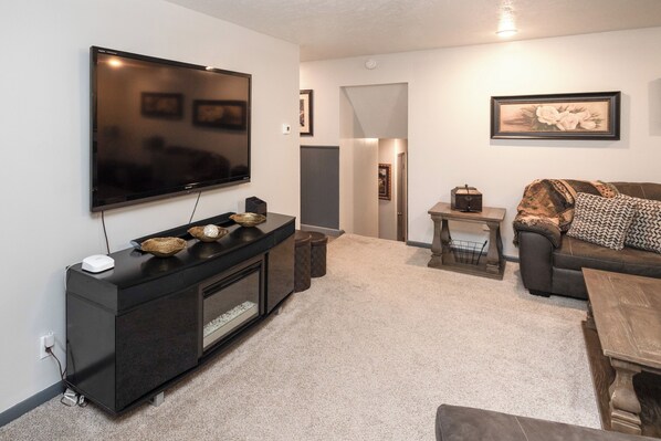 A living room with a flat-screen TV mounted on the wall, a dark entertainment unit, a brown sofa with patterned cushions, a small wooden coffee table, and a framed picture on the wall.